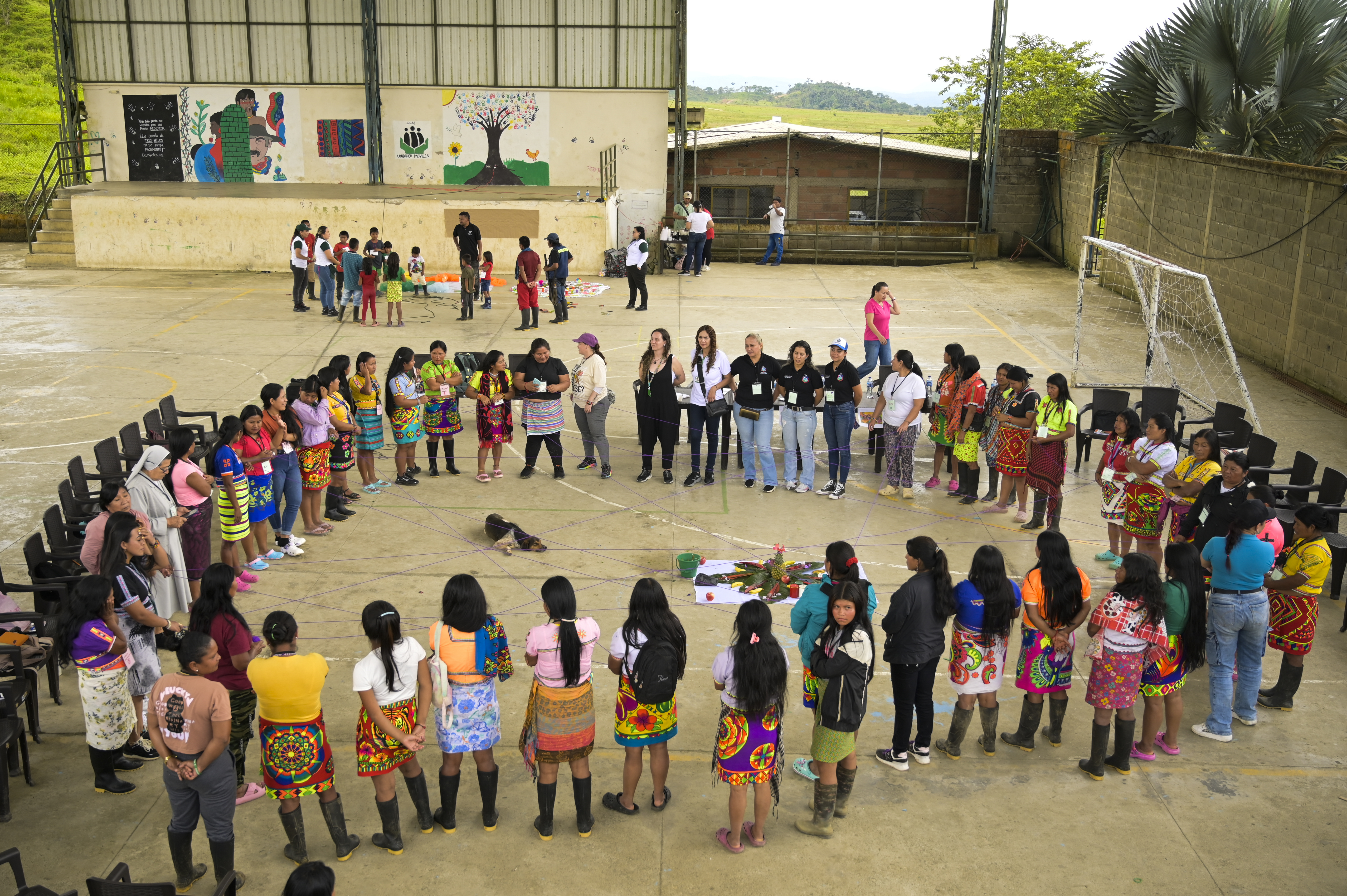 Grupo de mujeres y facilitadores reunidos en círculo en una cancha cubierta durante una actividad comunitaria, con vestimenta tradicional colorida y materiales en el centro.